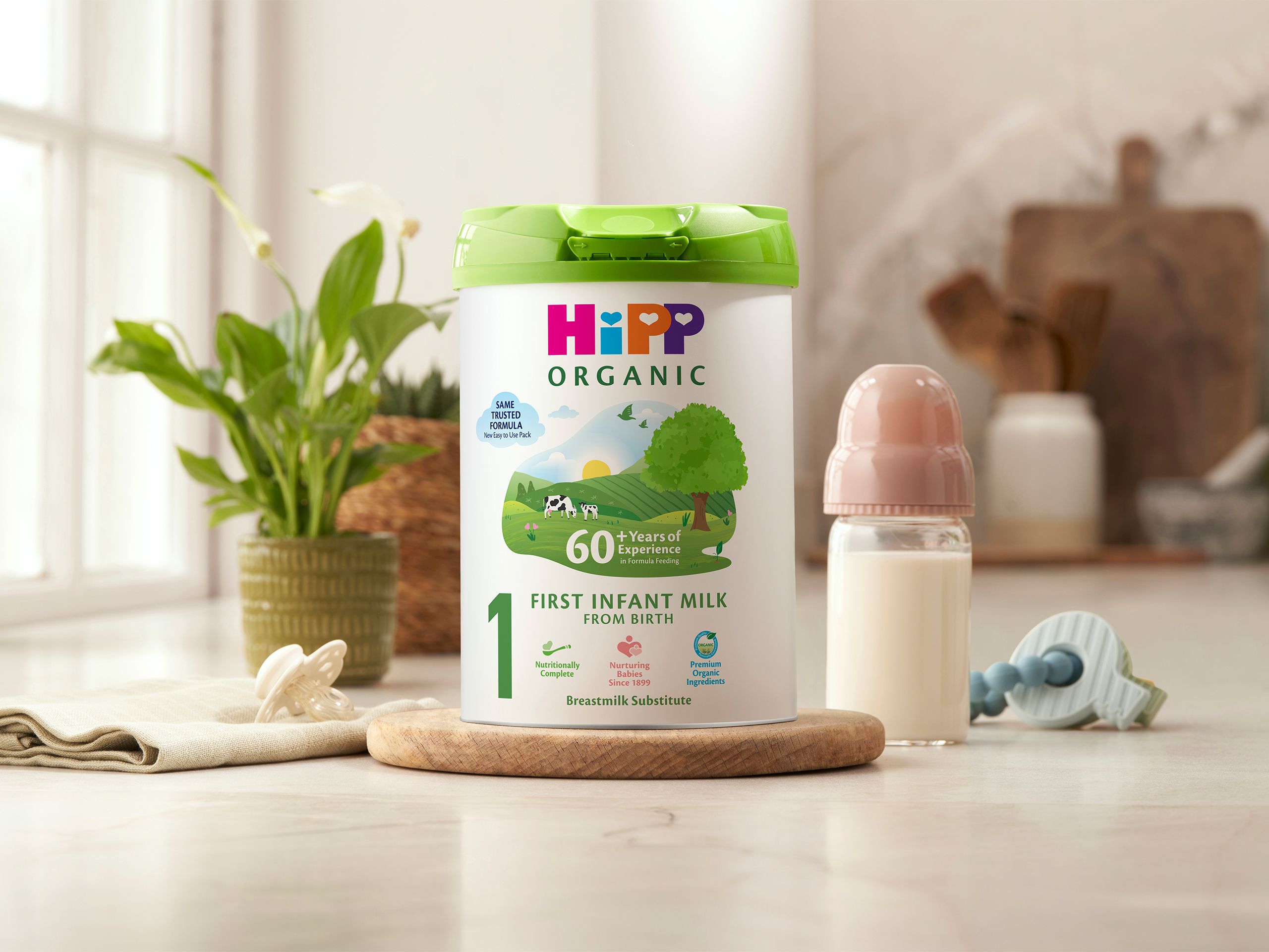 HiPP Organic First Infant Milk tin on a bright kitchen worktop beside a baby bottle, dummy and toy