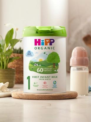 HiPP Organic First Infant Milk tin on a bright kitchen worktop beside a baby bottle, dummy and toy