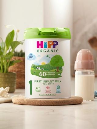 HiPP Organic First Infant Milk tin on a bright kitchen worktop beside a baby bottle, dummy and toy
