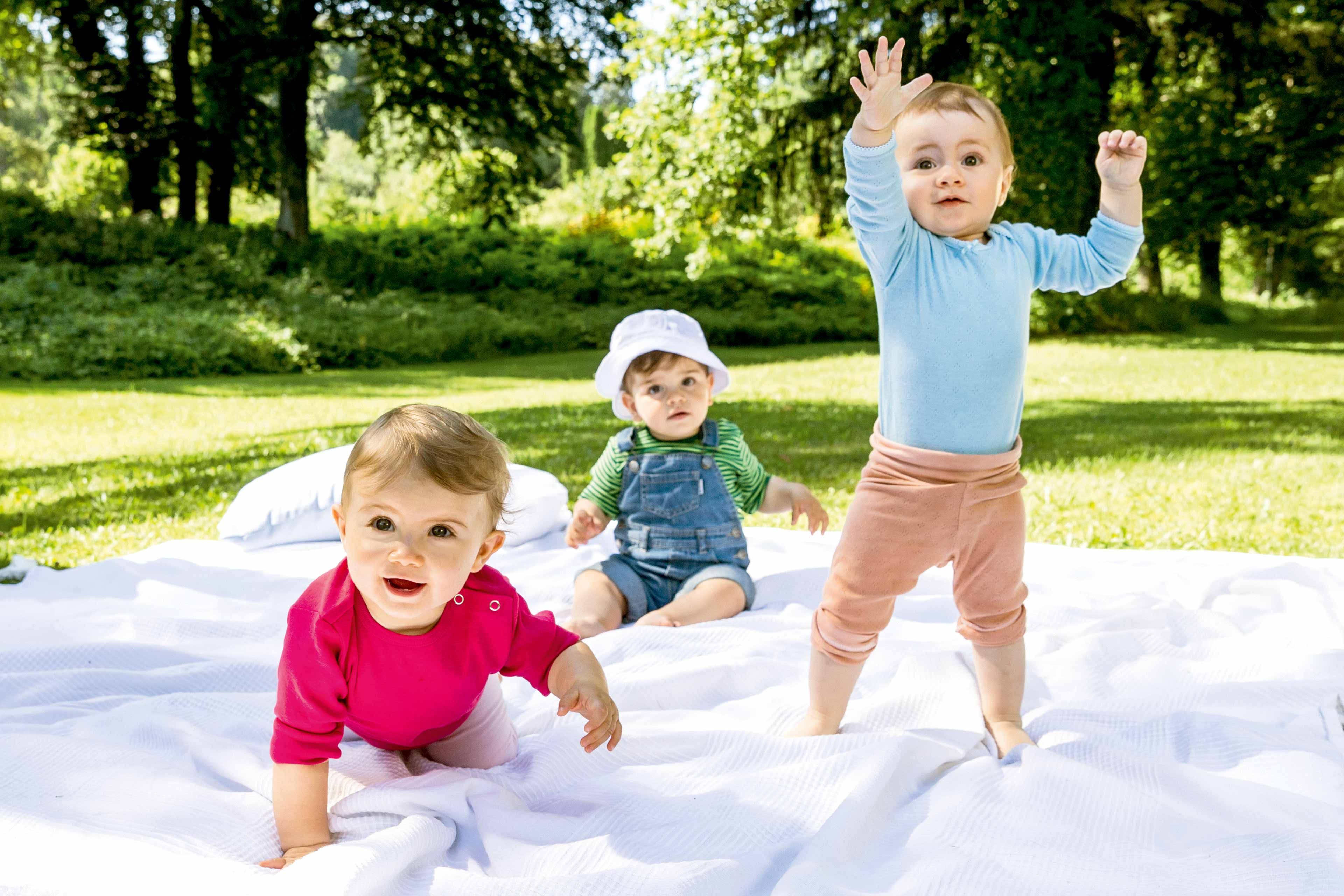  	Drei Kleinkinder spielen im Freien auf einer weißen Decke, umgeben von Grün.