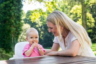 Ein kleines, rosa gekleidetes Baby kaut auf einem weißen Gegenstand, während eine Frau, die vermutlich die Mutter des Babys ist, es anlächelt.