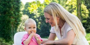 Ein kleines, rosa gekleidetes Baby kaut auf einem weißen Gegenstand, während eine Frau, die vermutlich die Mutter des Babys ist, es anlächelt.