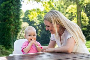 Ein kleines, rosa gekleidetes Baby kaut auf einem weißen Gegenstand, während eine Frau, die vermutlich die Mutter des Babys ist, es anlächelt.