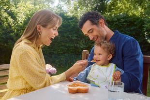 Eltern füttern ihr lachendes Baby im Garten mit Brei – gemeinsame Mahlzeit in vertrauter, liebevoller Atmosphäre.