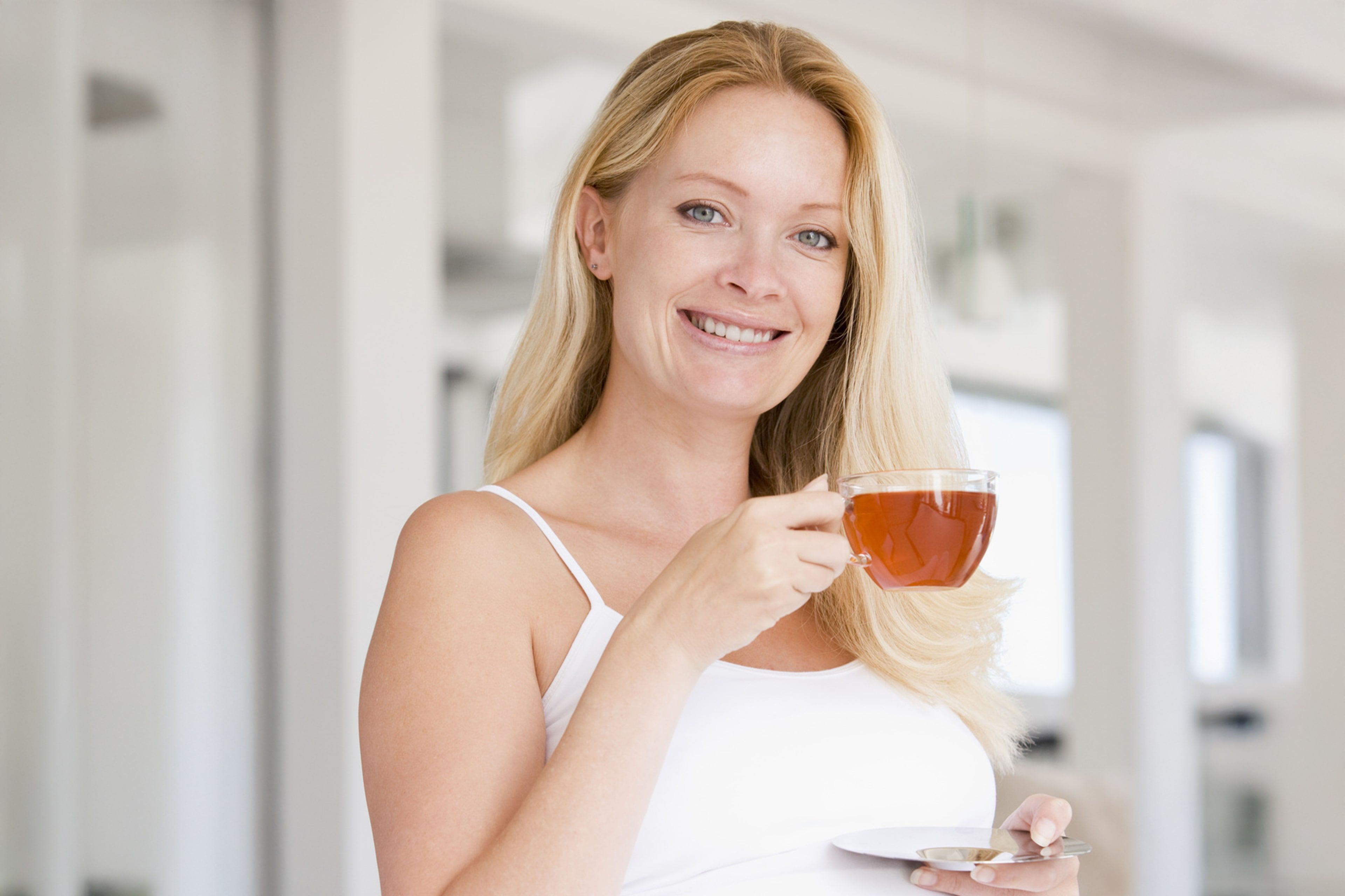 Eine Frau mit blondem Haar, die eine Tasse Tee in der Hand hält, steht an einem Fenster mit Blick ins Grüne.