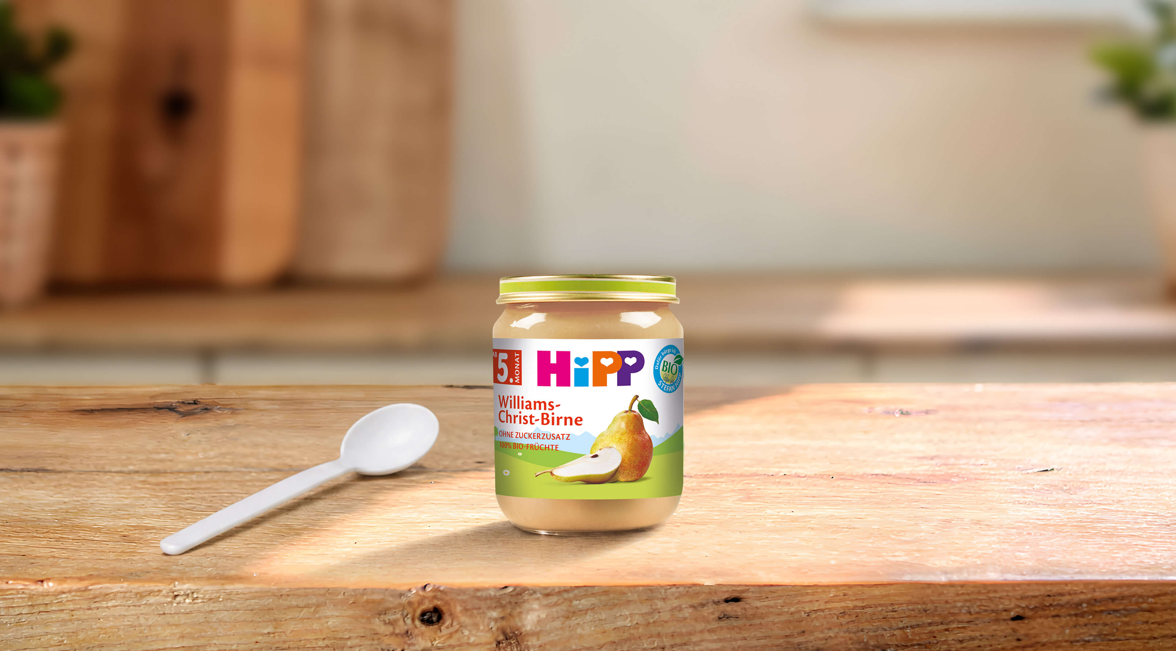 HiPP Bio Babygläschen Williams-Christ-Birne ab 5. Monat mit Löffel auf Holztisch in heller Küche