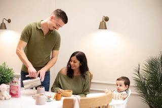 A man pours coffee into a mug while smiling at a woman seated at the table with a wooden bowl, both casually dressed and enjoying a cozy breakfast. A baby in a high chair looks on with curiosity, next to a box of HiPP Organic porridge and a soft toy on the table