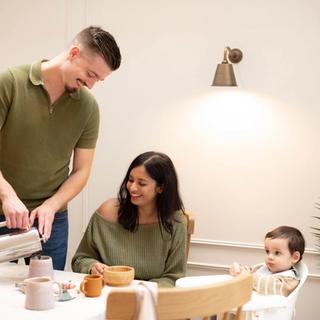 A man pours coffee into a mug while smiling at a woman seated at the table with a wooden bowl, both casually dressed and enjoying a cozy breakfast. A baby in a high chair looks on with curiosity, next to a box of HiPP Organic porridge and a soft toy on the table.