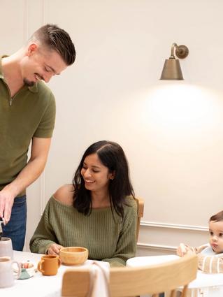 A man pours coffee into a mug while smiling at a woman seated at the table with a wooden bowl, both casually dressed and enjoying a cozy breakfast. A baby in a high chair looks on with curiosity, next to a box of HiPP Organic porridge and a soft toy on the table.