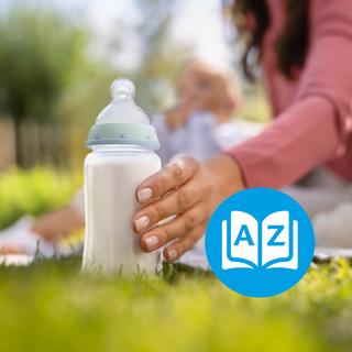 Elternhand greift im Grünen nach einer Babyflasche mit Milch, im Hintergrund liegt ein entspanntes Baby auf der Decke.