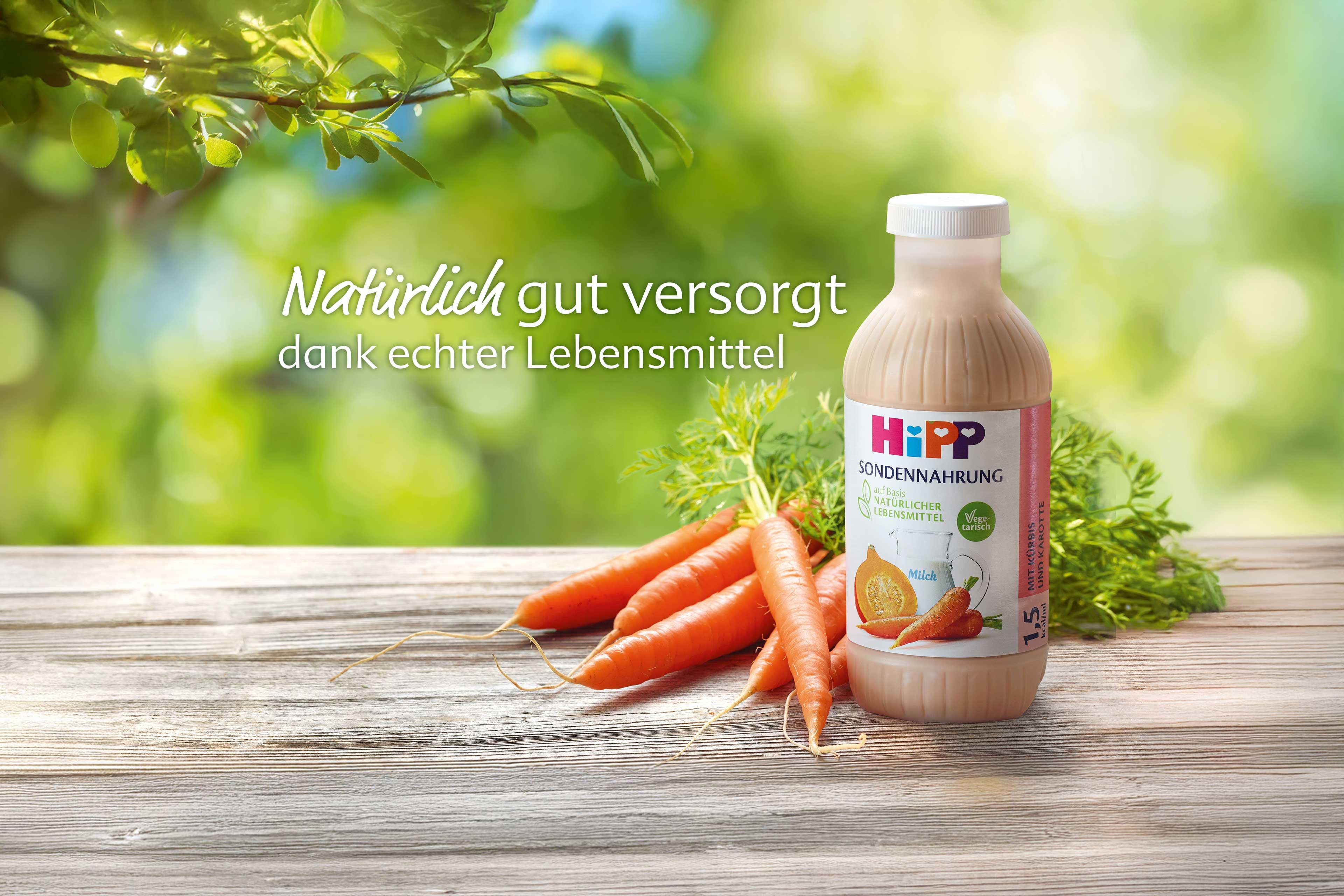 Frische Bundmöhren auf Holztisch vor grünem Hintergrund mit dem Schriftzug „Die natürlichste Art der künstlichen Ernährung“.
