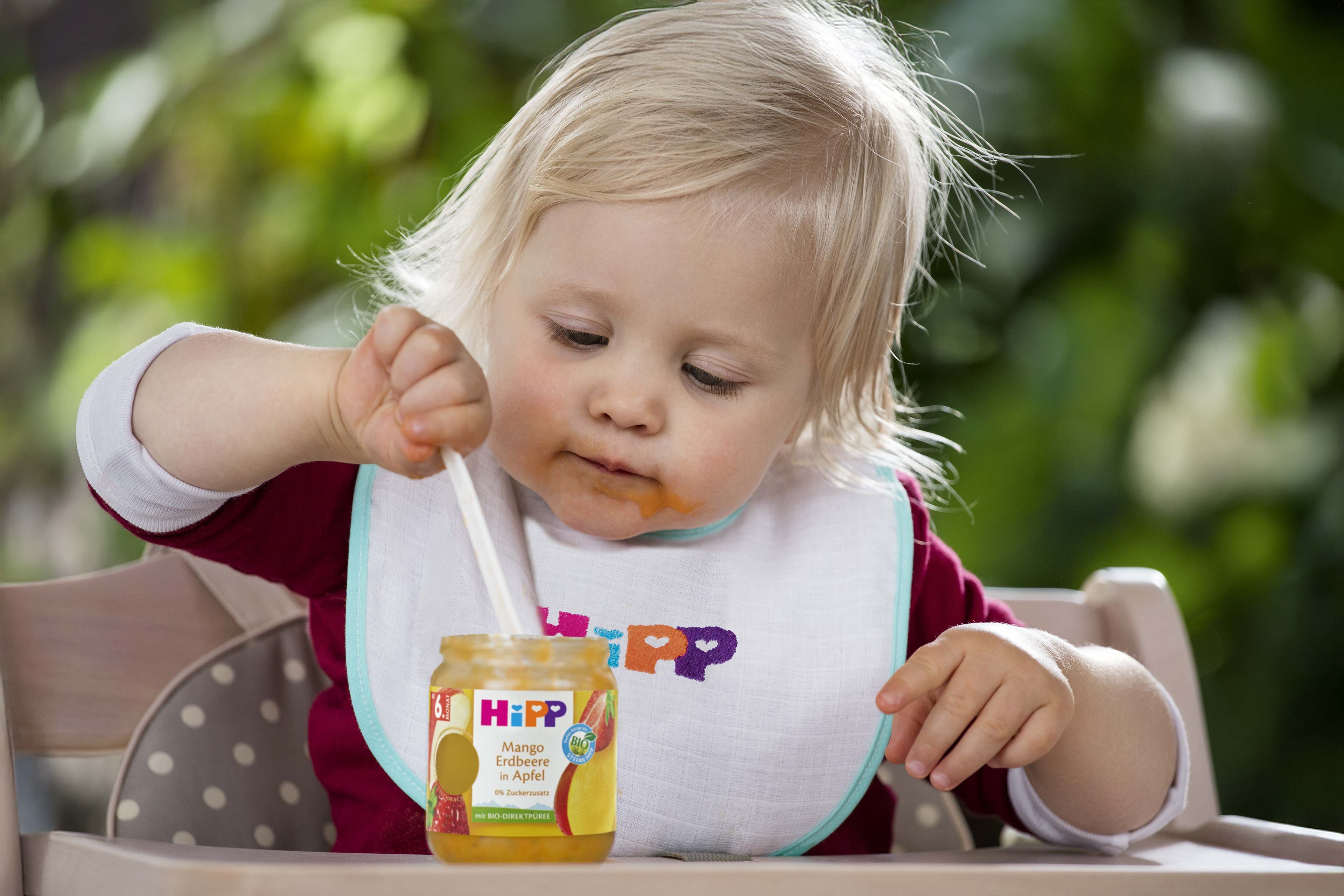 Kleinkind im Hochstuhl löffelt konzentriert HiPP Babybrei aus einem Glas mit Lätzchen um den Hals.