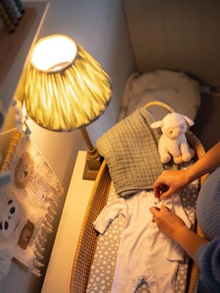 Pregnant woman standing beside a softly lit changing table, buttoning a white baby onesie next to a folded blanket and stuffed lamb in a cozy nursery.