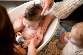 Baby in a bath being washed by parents, with another clothed baby sat next to them