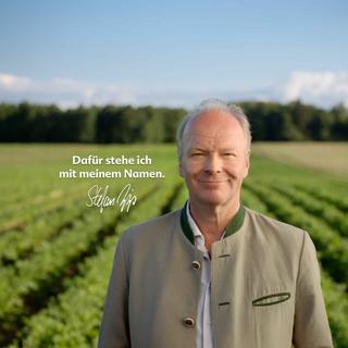 Lächelnder Mann in heller Trachtenjacke auf grünem Bio-Feld mit Schriftzug: „Dafür stehe ich mit meinem Namen.“