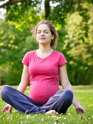 Eine Frau in einem rosa Hemd und einer blauen Hose sitzt in meditativer Haltung auf einer von Bäumen umgebenen Wiese.