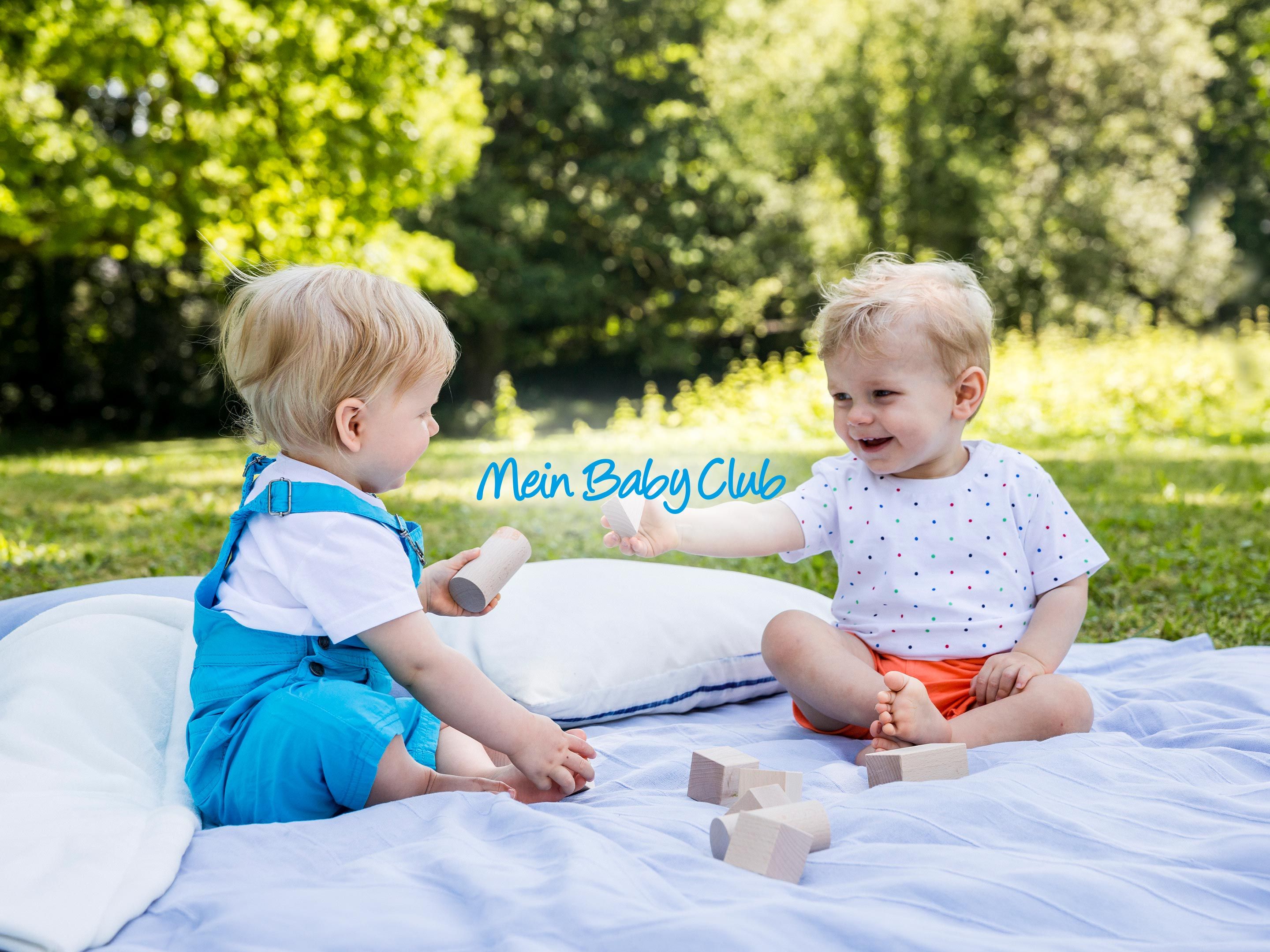 Zwei Kleinkinder spielen fröhlich mit Holzklötzen auf einer Decke im Grünen, mit dem Schriftzug „Mein BabyClub“ in der Mitte.