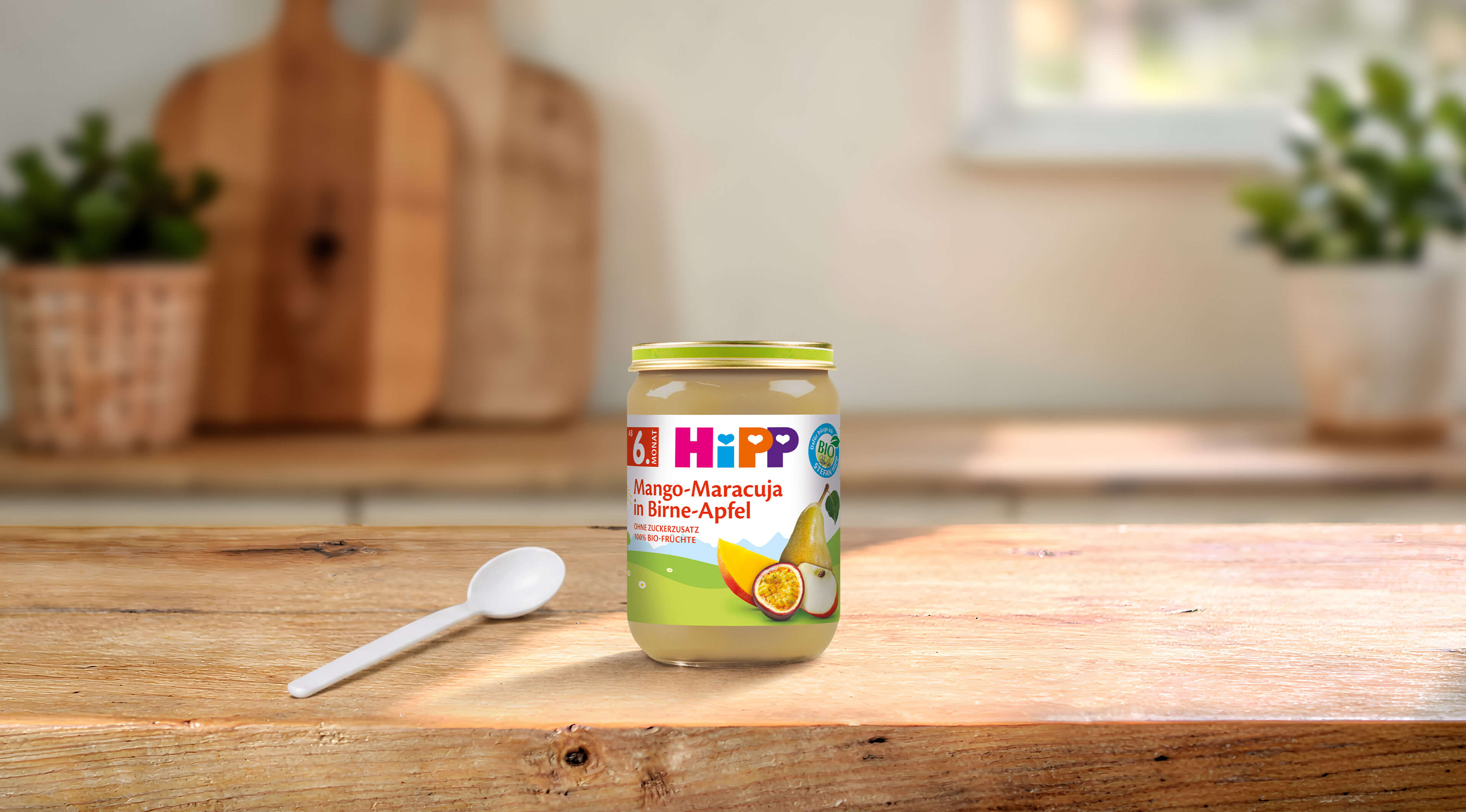HiPP Bio Fruchtpüree Mango-Maracuja in Birne-Apfel im Glas auf Holztisch mit Löffel in heller Küche
