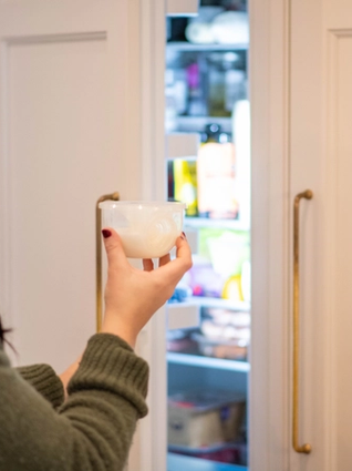 A woman with dark hair, wearing a green sweater, stands in front of an open refrigerator, holding a small white bowl. The refrigerator light illuminates various food items and condiments inside. The kitchen cabinets have gold handles, adding a touch of elegance to the scene.