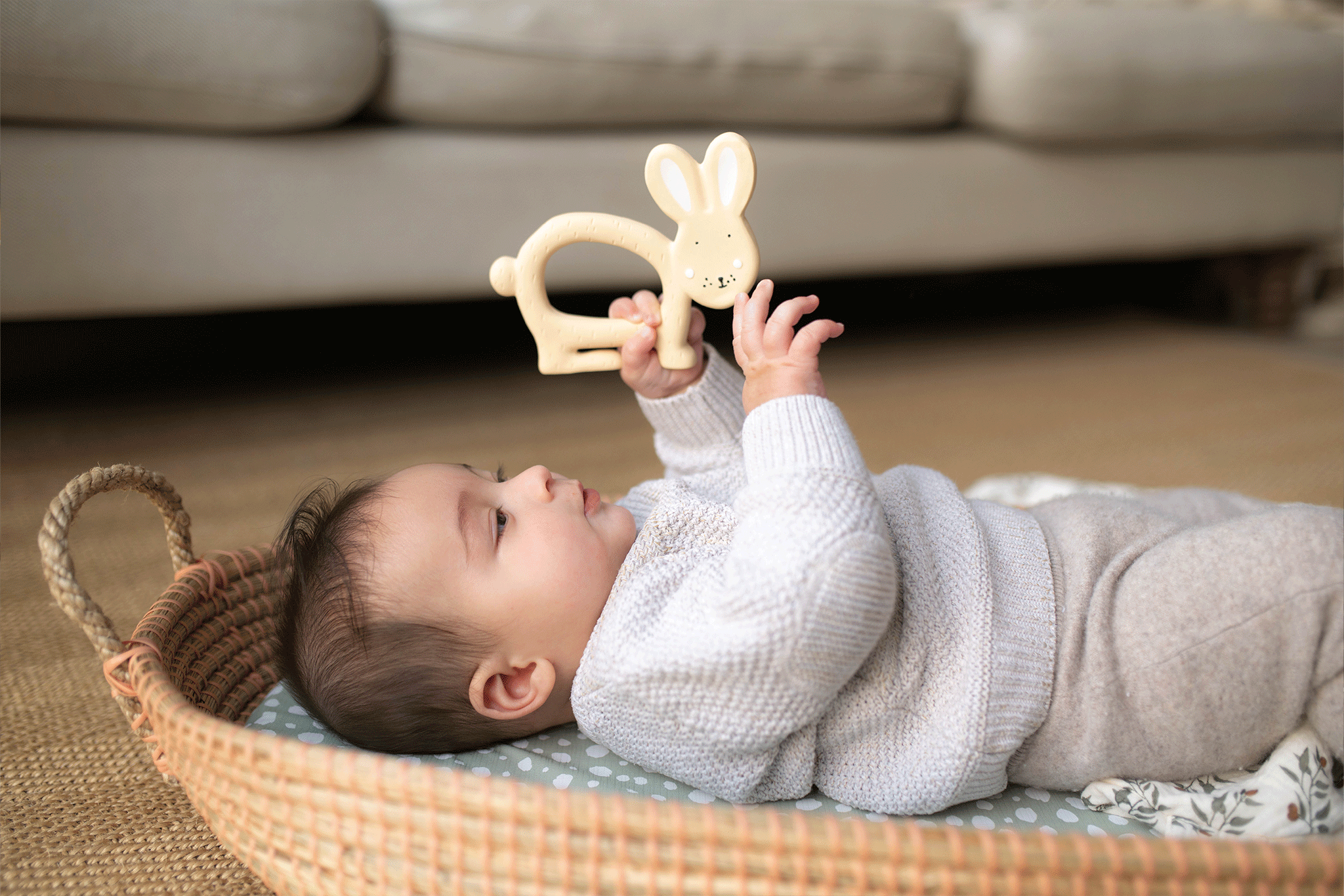 A baby dressed in a cream knitted jumper lying back in a seagrass Moses basket and playing with a cream rabbit teether.