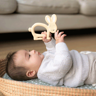 A baby dressed in a cream knitted jumper lying back in a seagrass Moses basket and playing with a cream rabbit teether.