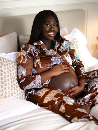 A pregnant woman sits comfortably on a bed, smiling warmly while cradling her bare belly. She’s wearing silky leopard-print pajamas, surrounded by soft pillows and warm, cozy lighting.