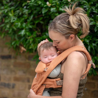 Parent cuddling a sleeping baby in a carrier outdoors, supporting calm contact napping and a soothing daytime sleep routine