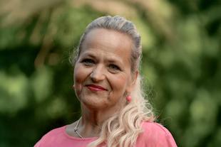Eine Frau mit blondem Haar, die ein rosa Oberteil und rosa Ohrringe trägt, vor einem grünen Hintergrund.