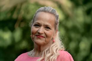 Eine Frau mit blondem Haar, die ein rosa Oberteil und rosa Ohrringe trägt, vor einem grünen Hintergrund.