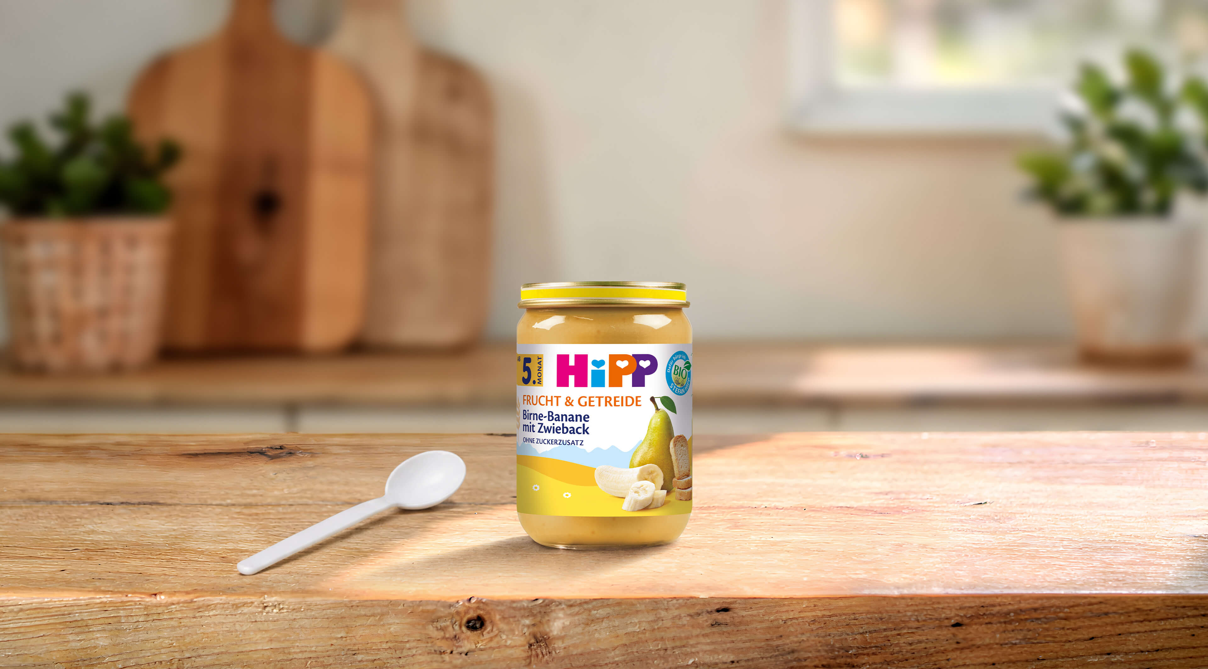 HiPP Bio-Gläschen Birne-Banane mit Zwieback, Frucht & Getreide ab 5. Monat auf Holztisch mit Löffel in heller Küche