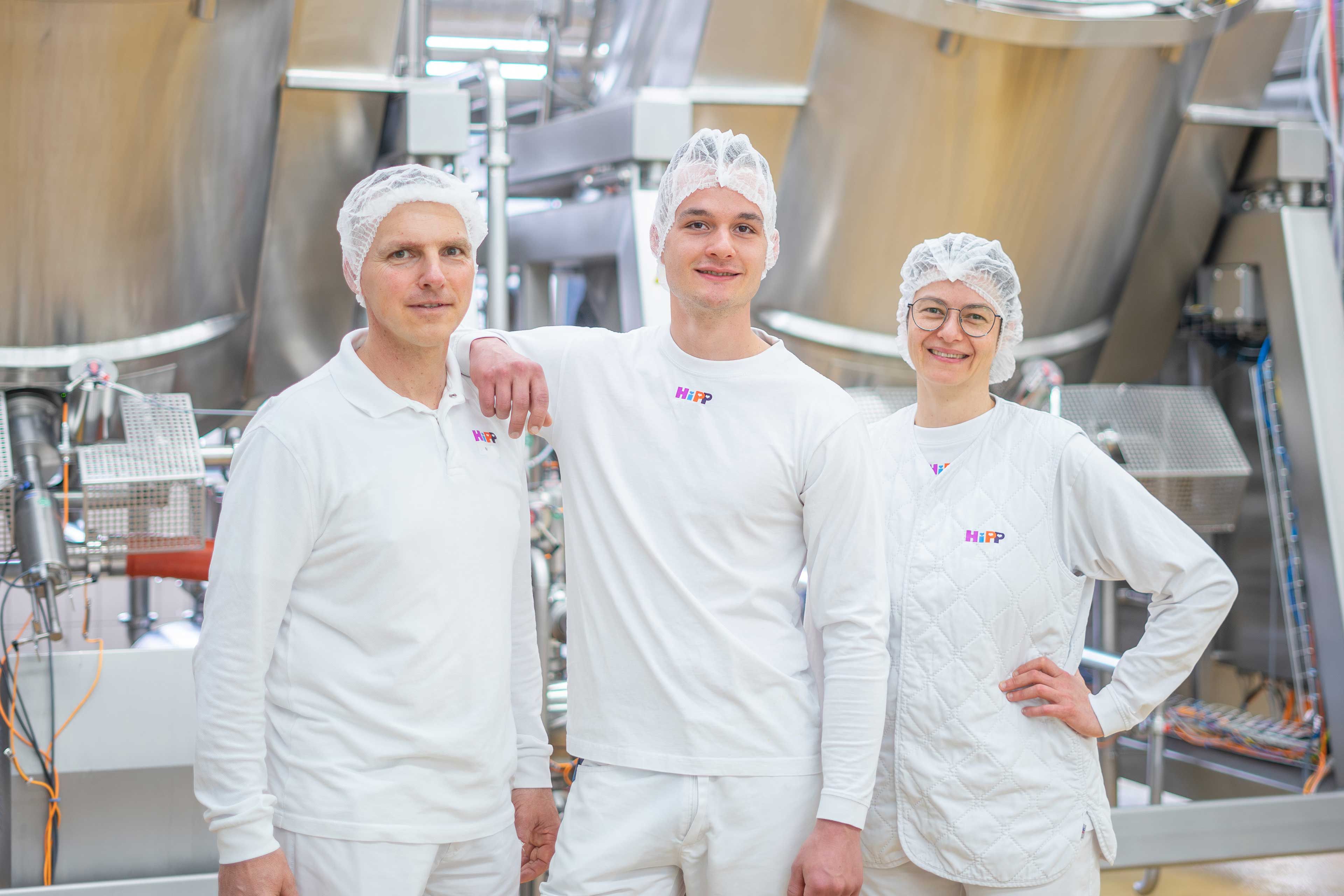 Drei HiPP Mitarbeitende in weißer Arbeitskleidung stehen lächelnd in einer sauberen Produktionsanlage zusammen