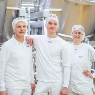 Drei HiPP Mitarbeitende in weißer Arbeitskleidung stehen lächelnd in einer sauberen Produktionsanlage zusammen