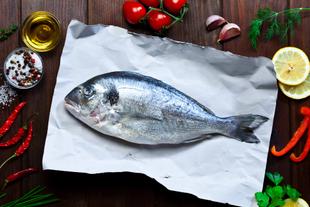 Ganze rohe Fischdorade auf Papier, umgeben von Zitronenscheiben, Kräutern, Öl, Salz und Gemüse als jodreiche Mahlzeit.