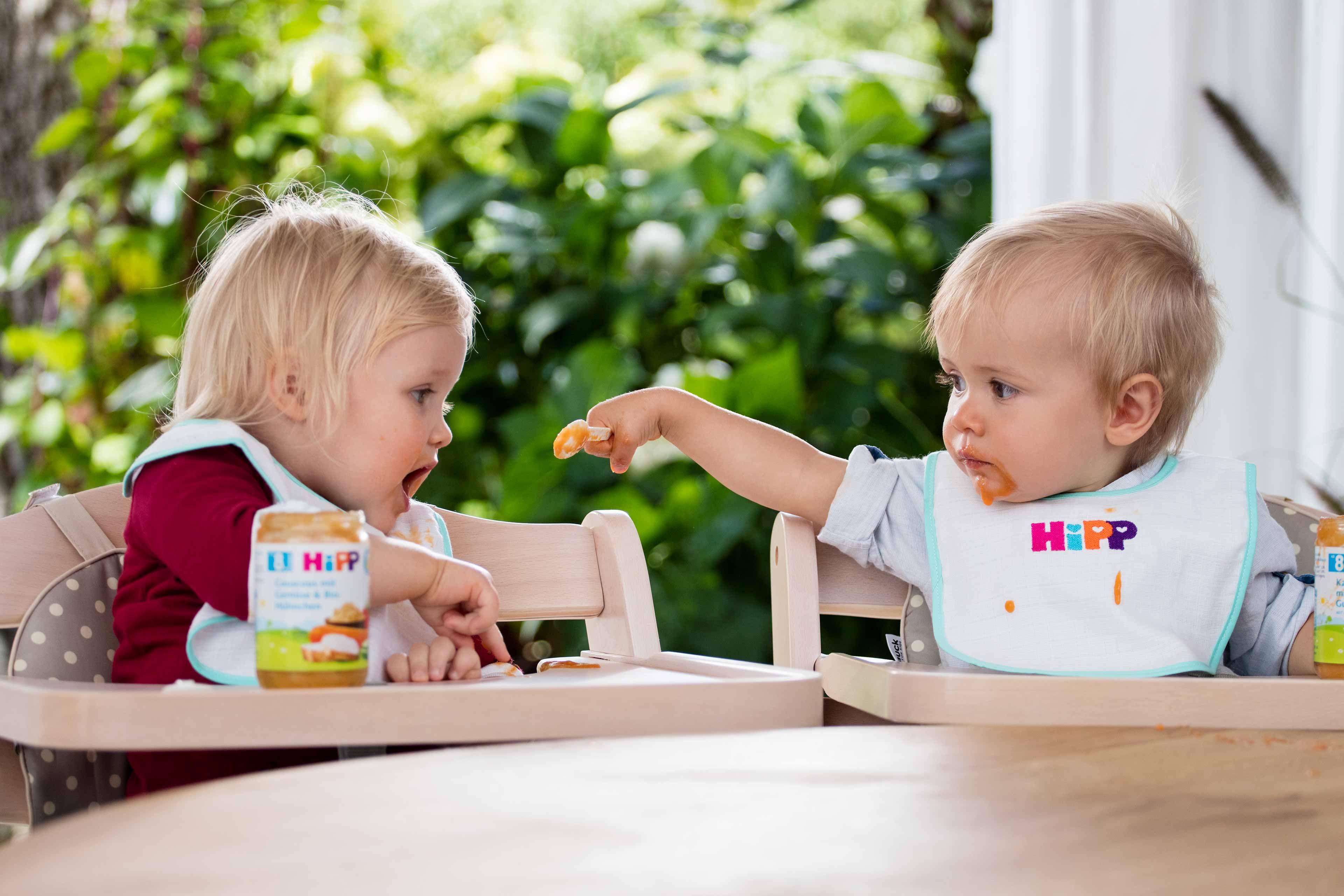 Zwei Kleinkinder, das eine füttert das andere mit einem Löffel, beide sitzen in Hochstühlen, umgeben von Gläsern mit Essen.