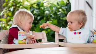 Zwei Kleinkinder, das eine füttert das andere mit einem Löffel, beide sitzen in Hochstühlen, umgeben von Gläsern mit Essen.