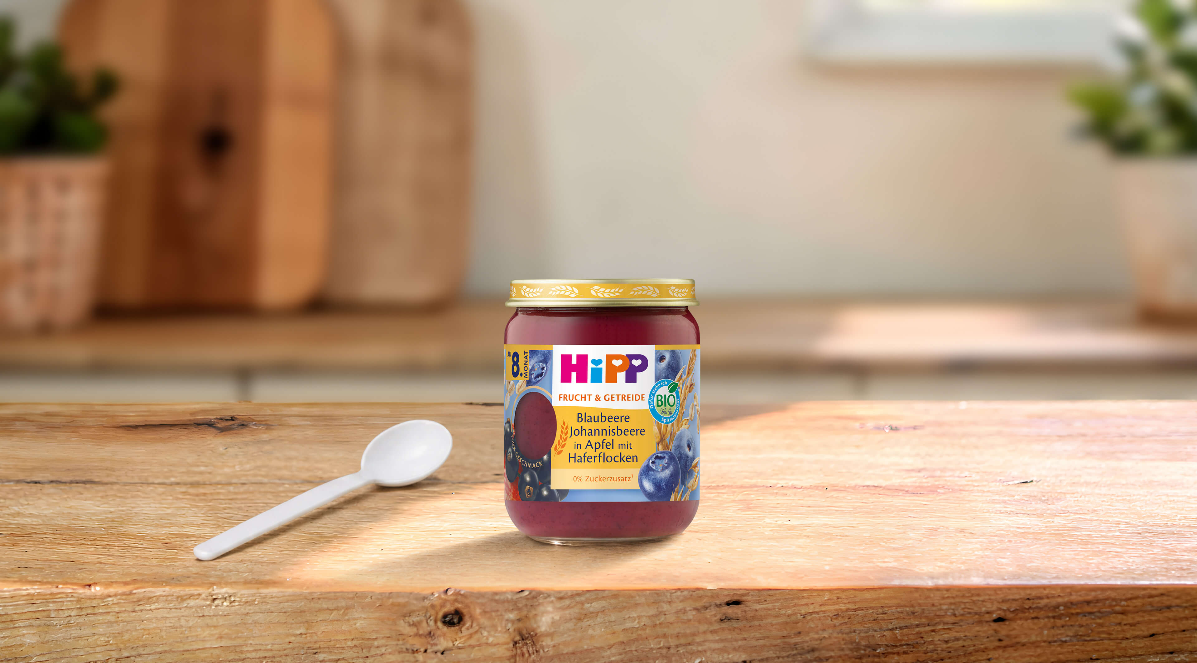 HiPP Bio-Gläschen Blaubeere Johannisbeere in Apfel mit Haferflocken auf Holztisch mit Löffel in heller Küche