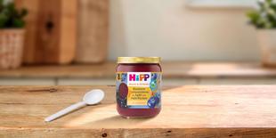 HiPP Bio-Gläschen Blaubeere Johannisbeere in Apfel mit Haferflocken auf Holztisch mit Löffel in heller Küche