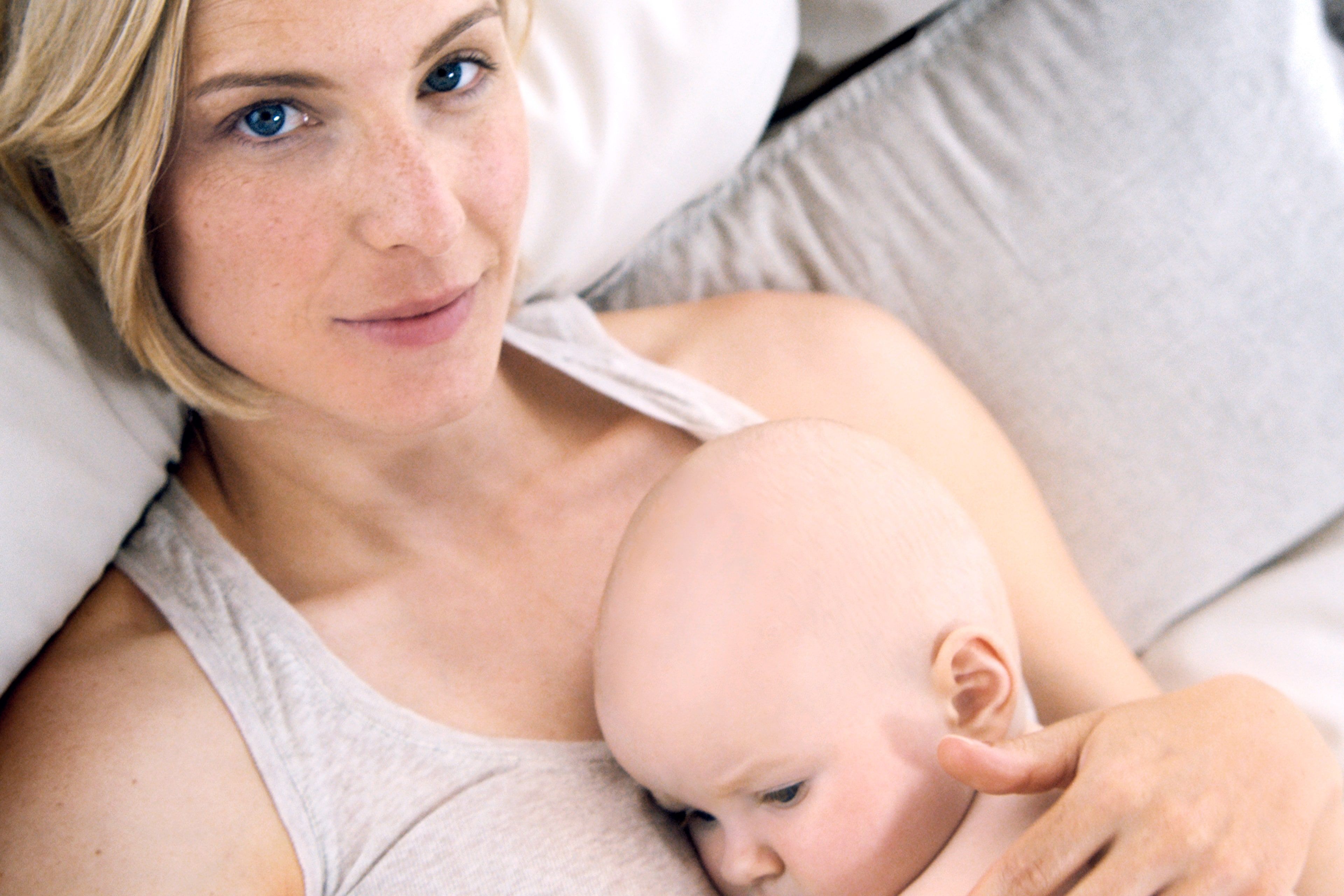 Eine Frau mit blondem Haar und blauen Augen, die ein Baby an sich drückt, das auf einer weichen Unterlage liegt.