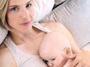 Eine Frau mit blondem Haar und blauen Augen, die ein Baby an sich drückt, das auf einer weichen Unterlage liegt.