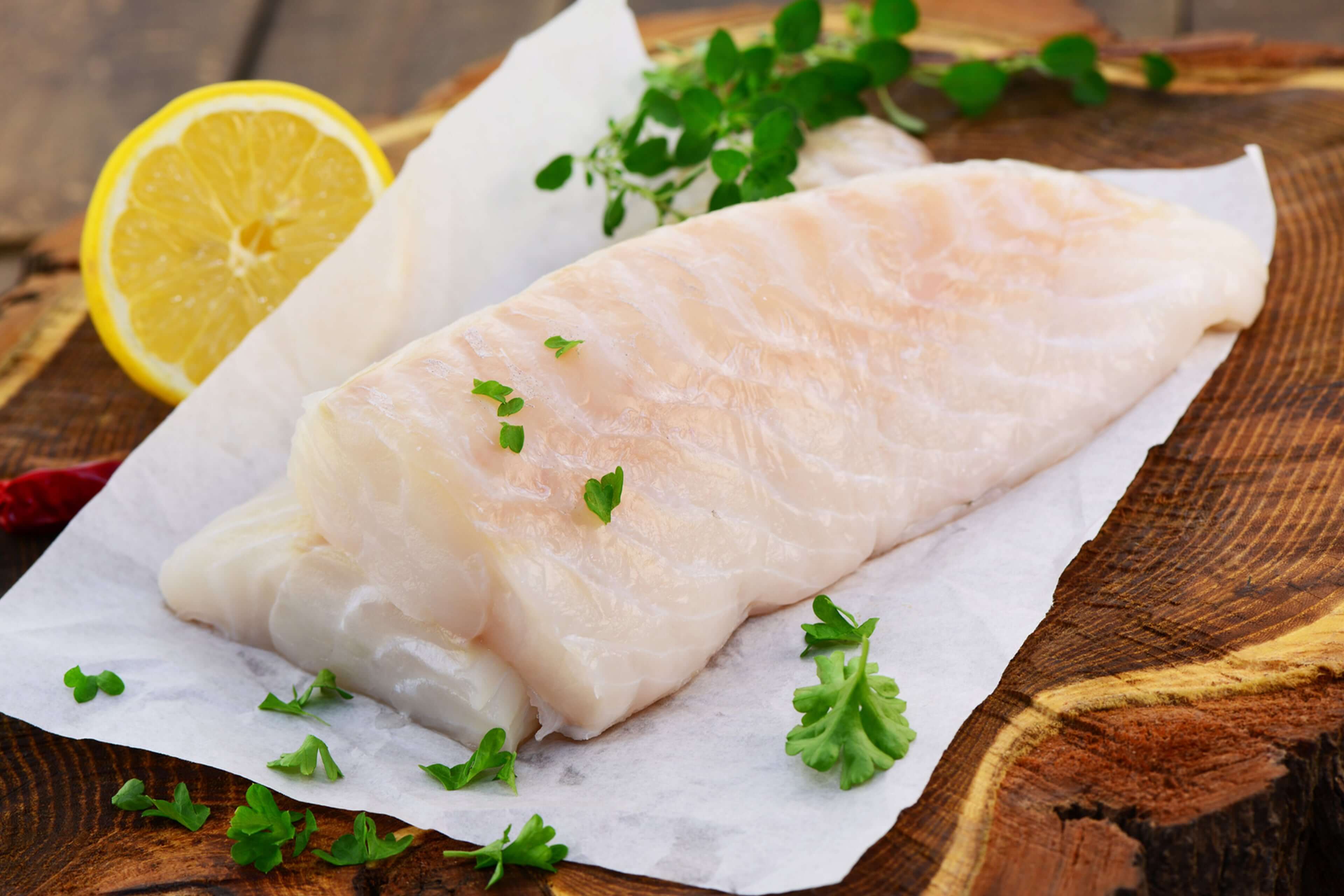 Ein Stück roher Fisch auf weißem Papier, garniert mit grünen Kräutern und einer Zitronenscheibe, auf einer Holzunterlage platziert.