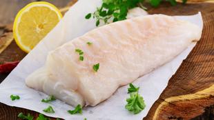 Ein Stück roher Fisch auf weißem Papier, garniert mit grünen Kräutern und einer Zitronenscheibe, auf einer Holzunterlage platziert.