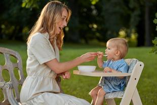 Mutter füttert ihr Baby im Hochstuhl mit Brei im Garten während der Beikosteinführung.