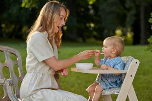 Mutter füttert ihr Baby im Hochstuhl mit Brei im Garten während der Beikosteinführung.