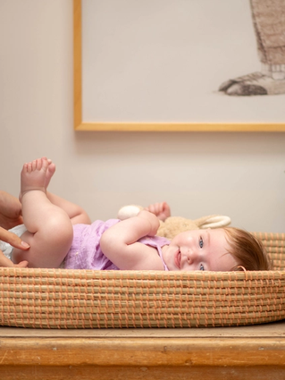 Mum changing baby in purple dress nappy, baby is smiling whilst holding her feet