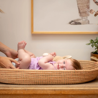 Mum changing baby in purple dress nappy, baby is smiling whilst holding her feet