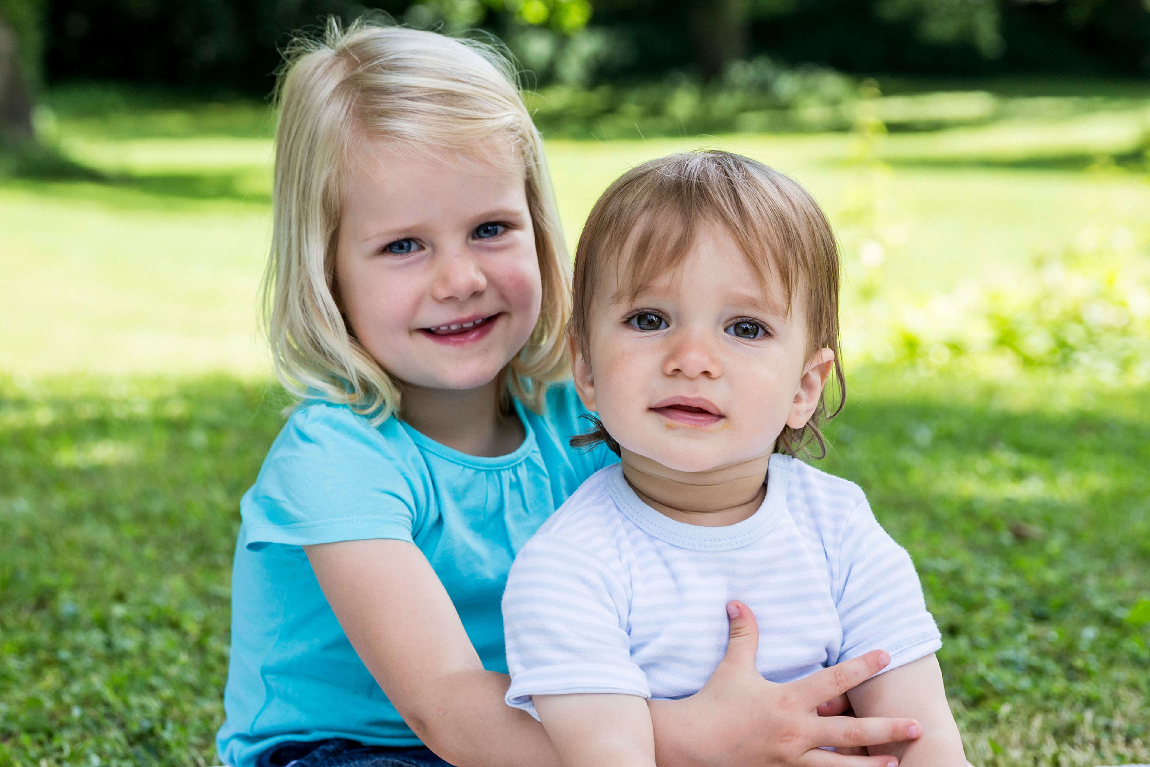 Zwei kleine Kinder, ein Mädchen und ein Junge, sitzen im Freien auf einer Wiese mit Bäumen im Hintergrund.
