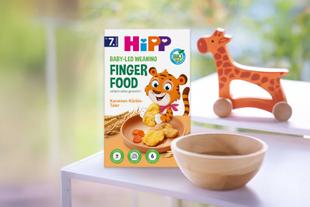 Packung Hipp Baby-led Weaning Finger Food mit Tiger-Illustration neben Holzschale und Holzgiraffe auf weißem Tisch