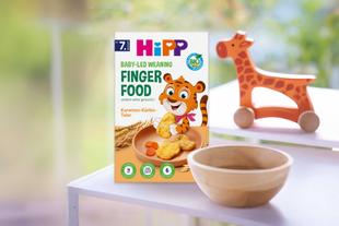 Packung Hipp Baby-led Weaning Finger Food mit Tiger-Illustration neben Holzschale und Holzgiraffe auf weißem Tisch