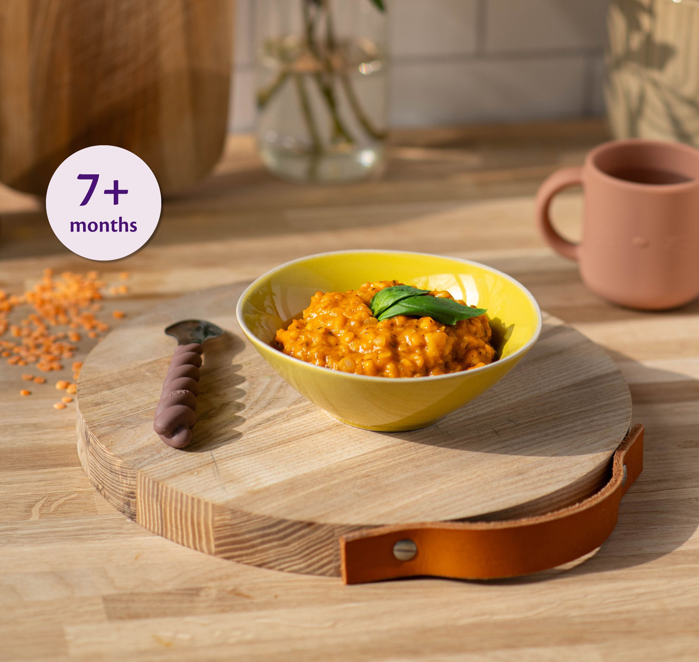 Bowl of soft lentil and tomato pasta for babies 7+ months on a wooden board with baby spoon and cup nearby