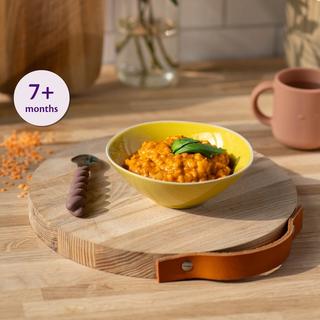 Bowl of soft lentil and tomato pasta for babies 7+ months on a wooden board with baby spoon and cup nearby